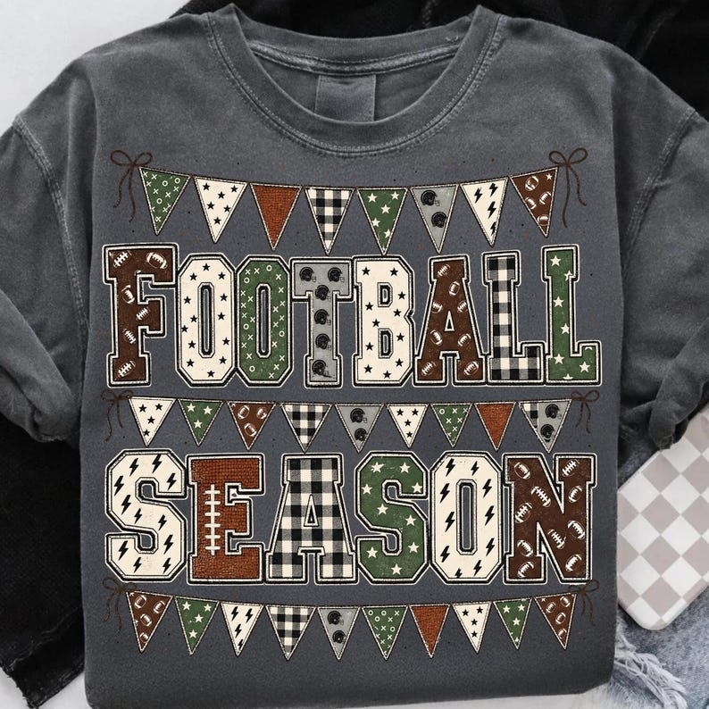 May include: A charcoal gray t-shirt featuring the text FOOTBALL SEASON in large block letters. The letters are filled with various patterns including footballs, plaid, and stars. Above and below the text are rows of triangular bunting flags.