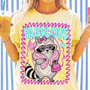 K&ouml;nnte beinhalten: Ein hellgelbes T-Shirt mit einer Grafik eines Waschb&auml;ren, der eine rosa Federboa, eine rosa Haarschleife und herzf&ouml;rmige Sonnenbrillen tr&auml;gt. Der Waschb&auml;r h&auml;lt eine rosa Handtasche. Der Text FABULOUS steht in Blau &uuml;ber dem Waschb&auml;ren, umgeben von Sternen.