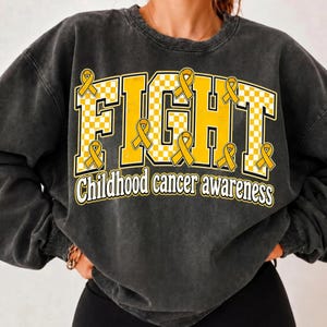 May include: A charcoal grey crewneck sweatshirt featuring the word FIGHT in large, yellow and white checkered block letters. Yellow awareness ribbons are integrated into the letters, with the text Childhood cancer awareness below in white.