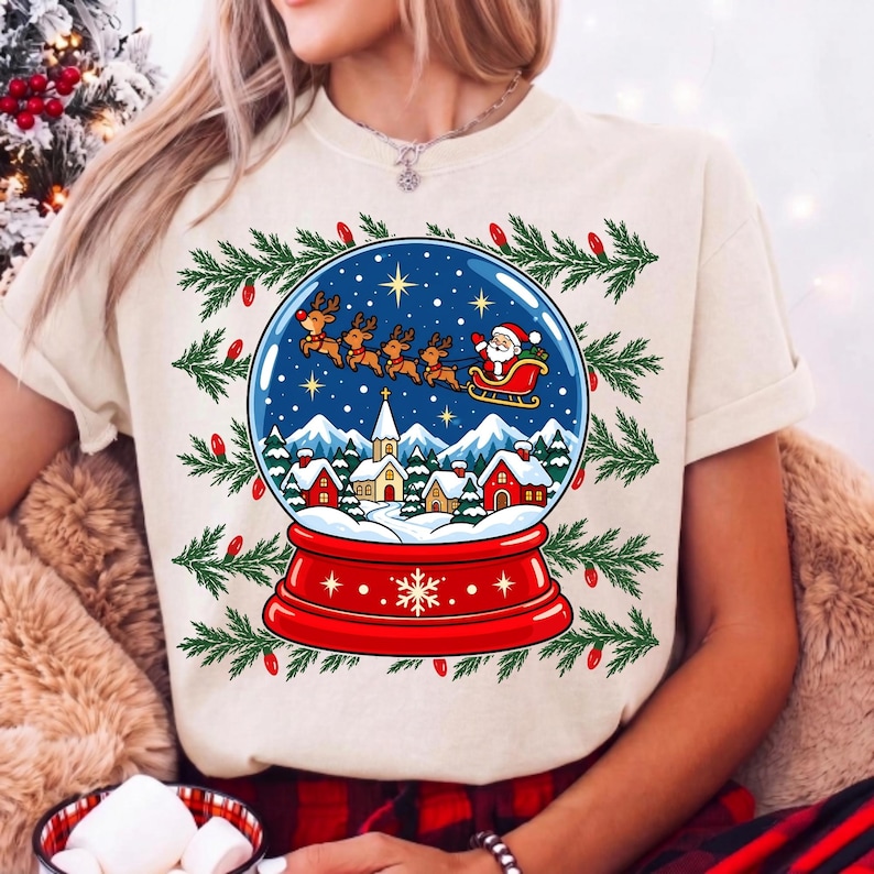May include: A cream-colored t-shirt featuring a graphic of a snow globe containing a winter village scene with houses, a church, and snow-capped mountains. Above the village, Santa Claus in a sleigh pulled by reindeer flies across a starry sky.