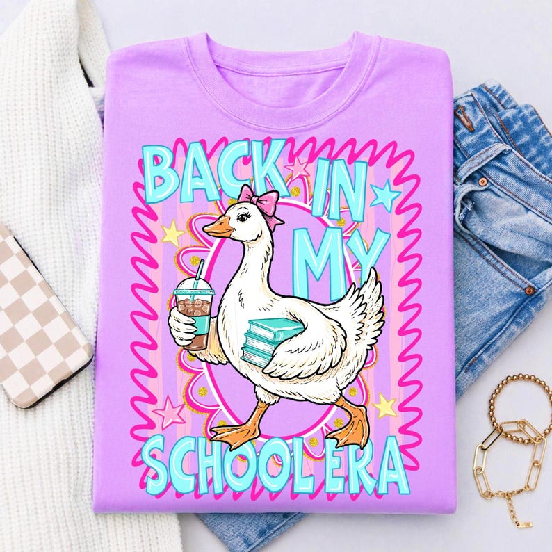 May include: A lilac-colored t-shirt with a graphic of a goose wearing a pink bow, holding a coffee and books. The text "BACK IN MY SCHOOL ERA" is in turquoise, surrounded by a pink and yellow design.