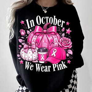 May include: A black sweatshirt featuring a graphic with the text In October We Wear Pink. The design includes a large pink pumpkin with a bow, a smaller polka-dot pumpkin, a coffee cup, a baseball cap, and roses, all accented with pink ribbons.