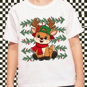 May include: A white t-shirt featuring a graphic of a cartoon reindeer wearing a green baseball cap and a red scarf. The reindeer is positioned in front of horizontal rows of green pine branches decorated with small red Christmas light bulbs.