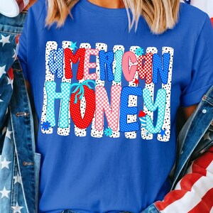 May include: A royal blue t-shirt featuring a graphic print with the text AMERICAN HONEY in a bold, multi-colored, polka-dot and checkered pattern. The design is set against a background of vertical white stripes with black dots and small stars.