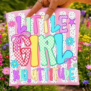 May include: A pink fabric panel featuring a white and black speckled stripe pattern. The text LITTLE GIRL BIG ATTITUDE is printed in large, colorful, checkered and polka-dot letters. The design includes pink bows and small colorful flowers.