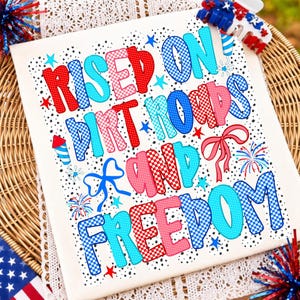 May include: A white t-shirt featuring the text "RAISED ON DIRT ROADS AND FREEDOM" in a bold, multi-coloured checkered pattern of red, light blue, and dark blue. The design includes small black dots, blue and red stars, fireworks, and bows.