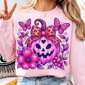 K&ouml;nnte beinhalten: Ein hellrosa Sweatshirt mit Rundhalsausschnitt und einem lebendigen Grafikdruck eines K&uuml;rbisgesichts mit einer Schleife im Leopardenmuster, umgeben von bunten Schmetterlingen, gro&szlig;en rosa Blumen und Beeren in Lila, Pink und Orange.
