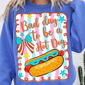 May include: A blue long-sleeve sweatshirt featuring a graphic print with a vertical pink, white, and blue striped background. The text reads Bad day to be a Hot Dog in orange. It includes a hot dog illustration, a patriotic bow, and yellow stars.