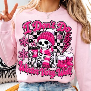 May include: A light pink sweatshirt featuring a graphic of a skeleton wearing a pink beanie and scarf, holding a pink coffee cup. The design includes a black and white checkered background, pink snowflakes, a leopard print lightning bolt, and the text I Don't Do Winter Very Well.
