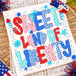 K&ouml;nnte beinhalten: Ein wei&szlig;es T-Shirt mit dem Text SWEET LAND OF LIBERTY in einer Mischung aus roten, wei&szlig;en und blauen Mustern, einschlie&szlig;lich Punkten und Karos. Das Design enth&auml;lt kleine blaue Blumen, hellblaue Sterne und rote und blaue Feuerwerksmotive.