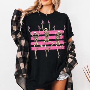 May include: A black t-shirt featuring a graphic of three dancing skeletons. Each skeleton holds a small pink ribbon above its head. The design includes three horizontal pink brushstroke stripes behind the skeletons, set against the black fabric.