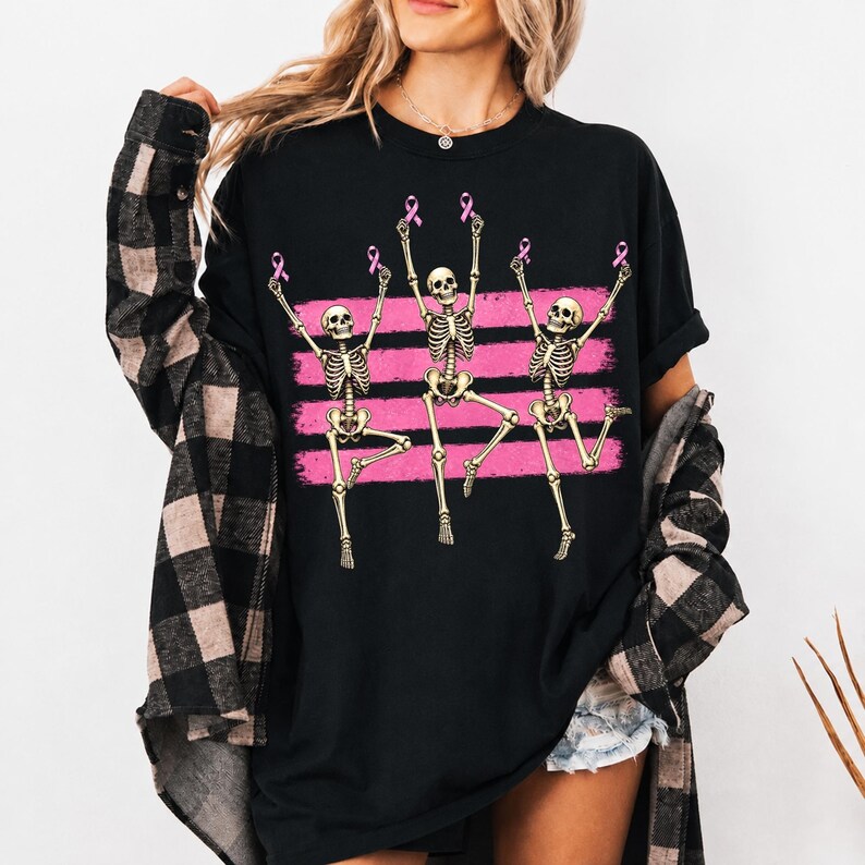 May include: A black t-shirt featuring a graphic of three dancing skeletons. Each skeleton holds a small pink ribbon above its head. The design includes three horizontal pink brushstroke stripes behind the skeletons, set against the black fabric.