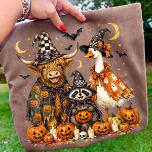 May include: A brown fabric tote bag featuring a graphic illustration of a Highland cow, a raccoon, and a goose wearing witch hats and Halloween costumes. The scene includes jack-o'-lanterns, ghosts, bats, stars, and crescent moons.