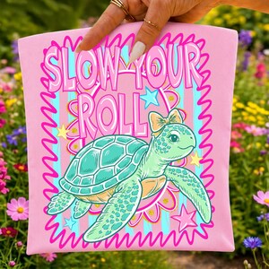 May include: A light pink fabric item featuring a graphic illustration of a green sea turtle wearing a small bow on its head. The text SLOW YOUR ROLL is printed in large pink letters above the turtle, surrounded by yellow stars and a pink border.