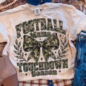 May include: An ivory short-sleeve t-shirt featuring a large graphic with the text "FOOTBALL MAMA TOUCHDOWN SEASON" in a green camouflage pattern. The design includes a central camouflage bow and leafy branches, accented by small star shapes.