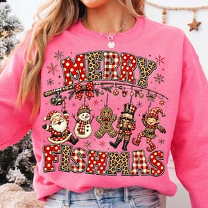 May include: A bright pink sweatshirt featuring a graphic print with the text MERRY FISHMAS. The design includes a fishing rod hanging ornaments shaped like Santa, a snowman, a gingerbread man, a nutcracker, and an elf, all with festive patterns.