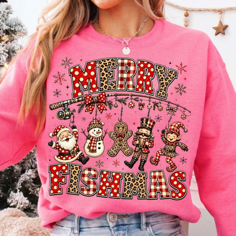 May include: A bright pink sweatshirt featuring a graphic print with the text MERRY FISHMAS. The design includes a fishing rod hanging ornaments shaped like Santa, a snowman, a gingerbread man, a nutcracker, and an elf, all with festive patterns.