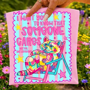 K&ouml;nnte beinhalten: Eine rosa Tragetasche mit einer bunten Grafik eines Waschb&auml;ren, der in einem Strandstuhl sitzt und ein Getr&auml;nk h&auml;lt. Der Text lautet I WANT YOU TO KNOW THAT SOMEONE CARES NOT ME, BUT SOMEONE. Das Design enth&auml;lt gelbe Sterne und einen Rand.