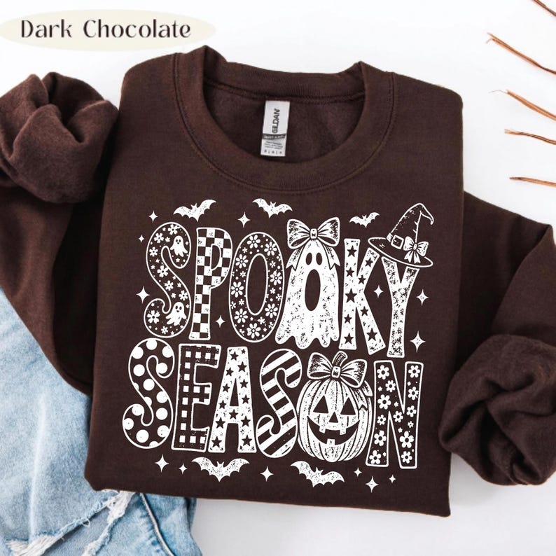 May include: A dark chocolate brown crewneck sweatshirt featuring a white graphic print that reads SPOOKY SEASON. The text incorporates patterns like checkers, stars, and polka dots, alongside illustrations of a ghost with a bow and a jack-o'-lantern.