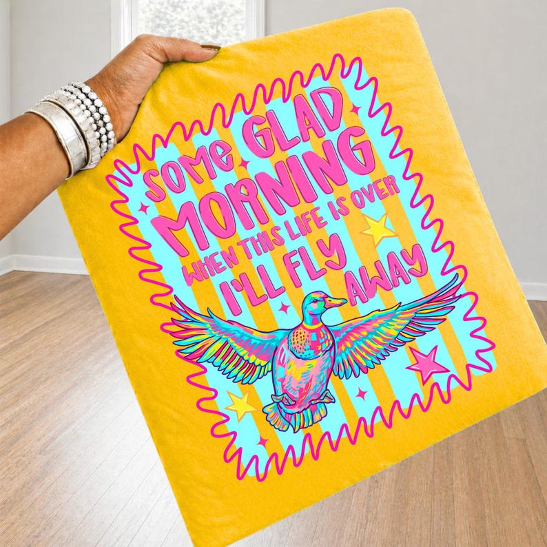 Pu&ograve; includere: Una federa quadrata gialla con una grafica vivace di un'anatra con le ali spiegate. Il testo recita SOME GLAD MORNING WHEN THIS LIFE IS OVER I'LL FLY AWAY. Il design include stelle e strisce nei colori rosa, blu e giallo.