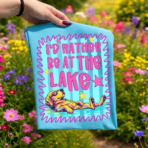 May include: A light blue t-shirt featuring a graphic of a dog floating in water with the text I'D RATHER BE AT THE LAKE in pink letters. The design includes a pink squiggly border, yellow stars, and a colorful dog wearing sunglasses.