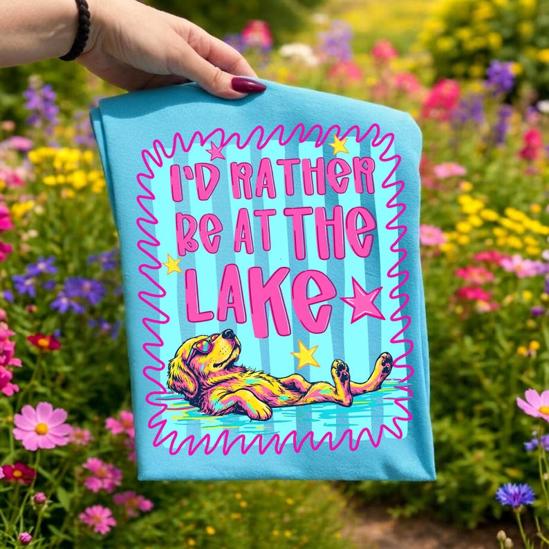 May include: A light blue t-shirt featuring a graphic of a dog floating in water with the text I'D RATHER BE AT THE LAKE in pink letters. The design includes a pink squiggly border, yellow stars, and a colorful dog wearing sunglasses.