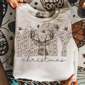 May include: A beige sweatshirt with the words "MERRY" in camouflage print, a deer graphic, and the word "christmas" in script. The design includes birds and stars. The sweatshirt is folded.