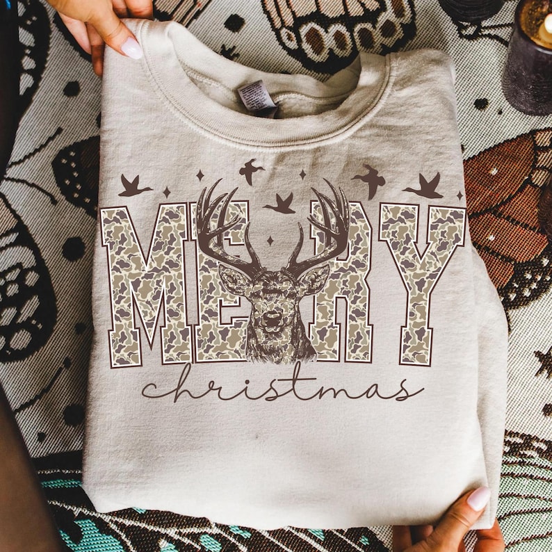May include: A beige sweatshirt with the words "MERRY" in camouflage print, a deer graphic, and the word "christmas" in script. The design includes birds and stars. The sweatshirt is folded.