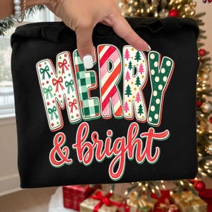 May include: A black sweatshirt featuring the text Merry & bright. The word Merry is in large block letters with varied patterns: red bows, green plaid, red and white candy cane stripes, pink and green trees, and green polka dots. Bright is in red.