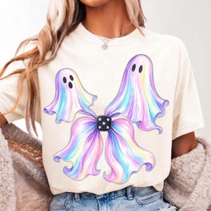 May include: A cream-coloured short-sleeve t-shirt featuring a graphic print of two ghosts with iridescent, pastel-coloured, flowing fabric-like bodies. The ghosts are positioned around a central black square die with white dots.