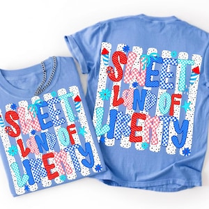 K&ouml;nnte beinhalten: Ein hellblaues T-Shirt mit einem Grafikdruck, der den Text SWEET LAND OF LIBERTY in rot-wei&szlig;-blauen Karomustern zeigt. Das Design umfasst Feuerwerksillustrationen, kleine blaue Blumen und Sterne auf einem Hintergrund aus vertikalen Streifen.