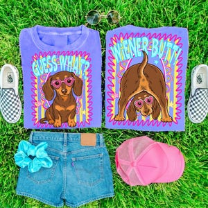 K&ouml;nnte beinhalten: Zwei violette T-Shirts mit einem braunen Dackel, der eine herzf&ouml;rmige Brille tr&auml;gt. Ein Shirt zeigt den Text "GUESS WHAT?", das andere "WIENER BUTT!". Ebenfalls enthalten sind blaue Jeansshorts, ein hellblaues Haargummi und eine rosa Netz-Baseballkappe.