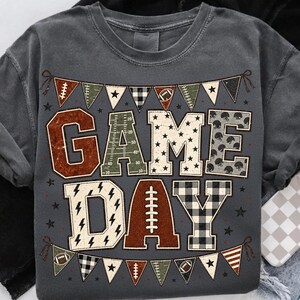 May include: A charcoal gray t-shirt featuring the text GAME DAY in large block letters. The letters are filled with various patterns including football field markings, stars, and plaid. A bunting banner with football motifs frames the text.