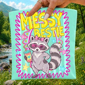May include: A light mint green tote bag featuring a graphic of a raccoon wearing pink heart-shaped glasses and a pink bow. The raccoon holds a drink with a heart icon. The text MESSY BESTIE is printed in yellow above the raccoon, surrounded by pink stars.