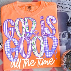 K&ouml;nnte beinhalten: Orangefarbenes Comfort Colors T-Shirt mit dem Schriftzug "GOD IS GOOD all the time" in einem Patchwork-Design. Die Buchstaben weisen Blumen-, Streifen- und Vichy-Muster mit lila Schleifen auf.