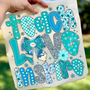 May include: A beige tote bag with the words "Teach Love Inspire" in turquoise, blue, and silver glitter. The design includes a bow with a snowman, snowflakes, and floral patterns. The bag is held by a hand.