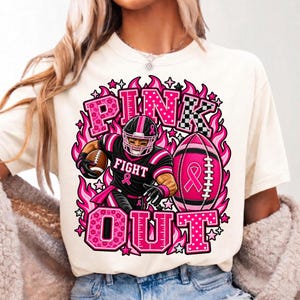 K&ouml;nnte beinhalten: Ein cremefarbenes T-Shirt mit einer Grafik eines Footballspielers in schwarzer Uniform mit rosa Akzenten. Der Text lautet PINK OUT. Ein rosa Football mit einem Schleifensymbol ist abgebildet, umgeben von rosa Flammen, Sternen und Punkten.