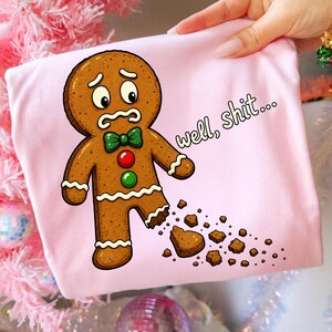 May include: A light pink t-shirt featuring a cartoon gingerbread person with a worried expression, a green bow tie, and red and green buttons. The gingerbread person has a broken leg with crumbs scattered below and the text well, shit...