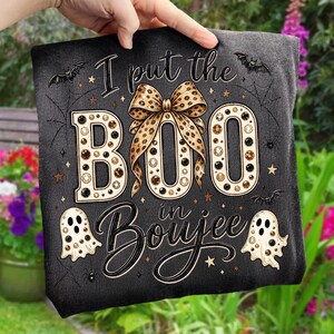 Pu&ograve; includere: Una t-shirt nera con il testo "I put the BOO in Boujee" in un carattere stilizzato. Il design include un grande fiocco con stampa leopardata, due fantasmi, piccoli pipistrelli, ragnatele e stelle, decorati con accenti circolari marroni e neri.
