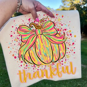 May include: A cream-colored t-shirt featuring a graphic of a pumpkin adorned with a large, multi-colored bow. The pumpkin design uses bright yellow, pink, and teal brushstrokes with gold glitter accents. The word thankful is printed below in yellow.