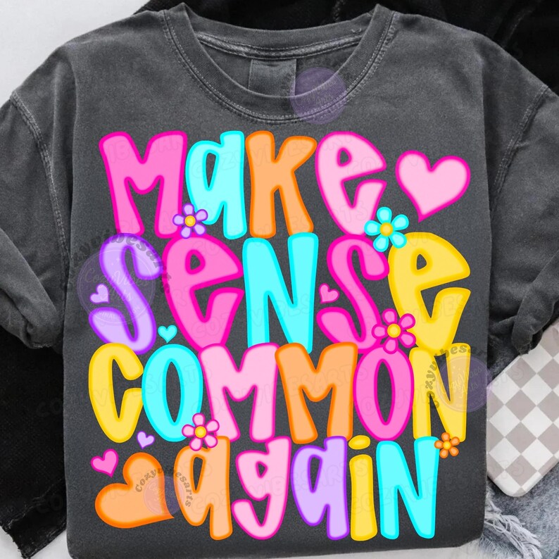 May include: Dark gray t-shirt with the phrase "MAKE SENSE COMMON AGAIN" in colorful bubble letters. The design includes hearts and flowers in pink, orange, yellow, and blue.