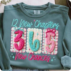 Puede incluir: Sudadera de color turquesa con texto bordado que dice "12 New Chapters" y "365 New Chances". Los n&uacute;meros "365" est&aacute;n en un estilo de ganchillo, con colores rosa y turquesa. La sudadera tiene cuello redondo y mangas largas.