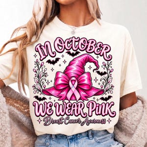 May include: A cream-colored t-shirt featuring a graphic of a bright pink witch hat adorned with a pink ribbon. The text reads "IN OCTOBER WE WEAR PINK" and "Breast Cancer Awareness," surrounded by black bats, stars, and decorative floral vines.