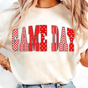 May include: Cream-colored sweatshirt with the words "GAME DAY" in red. The letters are filled with various patterns, including checkered, houndstooth, stars, stripes, and chevrons. A casual top for sports fans.