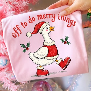 May include: A light pink t-shirt featuring a graphic of a white goose wearing a red Santa hat, a red vest with white trim, and red boots. The text "off to do merry things" is printed in red above the goose, accompanied by two holly sprigs.