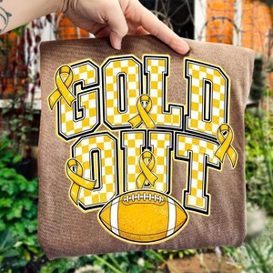 Puede incluir: Una camiseta marr&oacute;n con el texto GOLD OUT en un patr&oacute;n de cuadros amarillos y blancos. El dise&ntilde;o incluye varios lazos amarillos de concienciaci&oacute;n y un bal&oacute;n de f&uacute;tbol americano amarillo con rayas blancas bajo el texto, todo con borde negro.