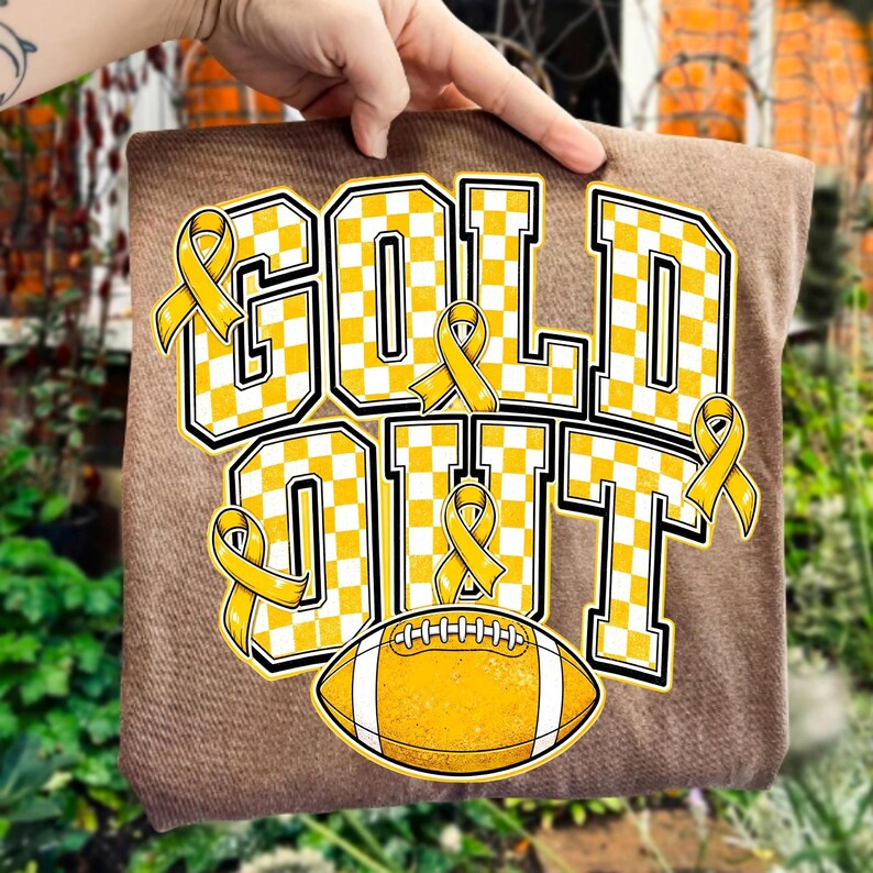Puede incluir: Una camiseta marr&oacute;n con el texto GOLD OUT en un patr&oacute;n de cuadros amarillos y blancos. El dise&ntilde;o incluye varios lazos amarillos de concienciaci&oacute;n y un bal&oacute;n de f&uacute;tbol americano amarillo con rayas blancas bajo el texto, todo con borde negro.