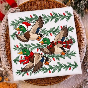 May include: A white t-shirt featuring a graphic print of three flying mallard ducks. One duck wears a red Santa hat, one wears a red and green plaid scarf, and one wears a red bow. The background pattern consists of green pine branches and red lights.