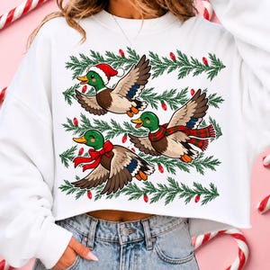 May include: A white long-sleeve sweatshirt featuring a graphic print of three mallard ducks in flight. The ducks are decorated with a red Santa hat, a red bow, and a red and green plaid scarf, surrounded by green pine branches and red light bulbs.