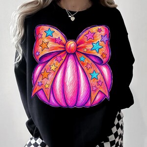 May include: A black long-sleeve sweatshirt featuring a large, vibrant graphic of a pumpkin decorated with a prominent bow. The design uses a scribbled, multi-colored aesthetic in shades of bright pink, orange, and blue, accented with star shapes.